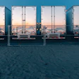 Row of industrial shipping containers at a waterfront dock with city skyline visible through open doors at sunset