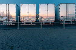 Row of industrial shipping containers at a waterfront dock with city skyline visible through open doors at sunset