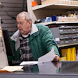 Older man in green jacket examining material samples at hardware store counter with shelves of products behind him