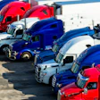 Fleet of red and blue semi-trucks lined up at a transportation depot with chrome details and industrial buildings in background