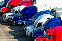 Fleet of red and blue semi-trucks lined up at a transportation depot with chrome details and industrial buildings in background
