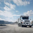 White Isuzu dump truck driving on dirt road at industrial quarry site with mountains and concrete plant in background