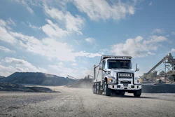 White Isuzu dump truck driving on dirt road at industrial quarry site with mountains and concrete plant in background