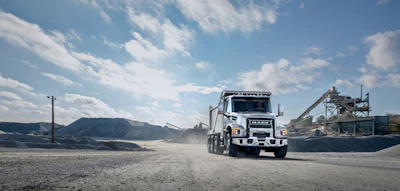 White Isuzu dump truck driving on dirt road at industrial quarry site with mountains and concrete plant in background