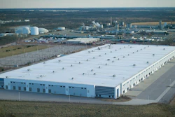 Aerial view of large industrial warehouse facility with white roof and blue structure surrounded by manufacturing plants and rural landscape