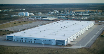Aerial view of large industrial warehouse facility with white roof and blue structure surrounded by manufacturing plants and rural landscape
