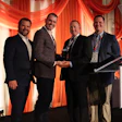 Four men in business suits stand together at NTDA event podium with red stage lighting, one holding an award