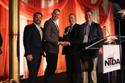Four men in business suits stand together at NTDA event podium with red stage lighting, one holding an award