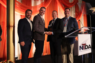 Four men in business suits stand together at NTDA event podium with red stage lighting, one holding an award