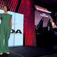 Woman in green jumpsuit presenting on stage at conference with large display screens showing branding and presentation content