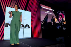 Woman in green jumpsuit presenting on stage at conference with large display screens showing branding and presentation content