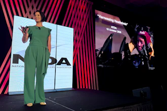 Woman in green jumpsuit presenting on stage at conference with large display screens showing branding and presentation content