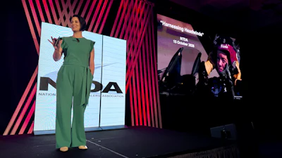 Woman in green jumpsuit presenting on stage at conference with large display screens showing branding and presentation content