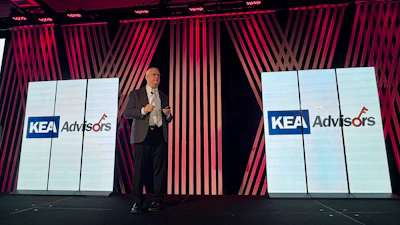Professional speaker presenting at corporate event with KEA Advisors branding displayed on large screens
