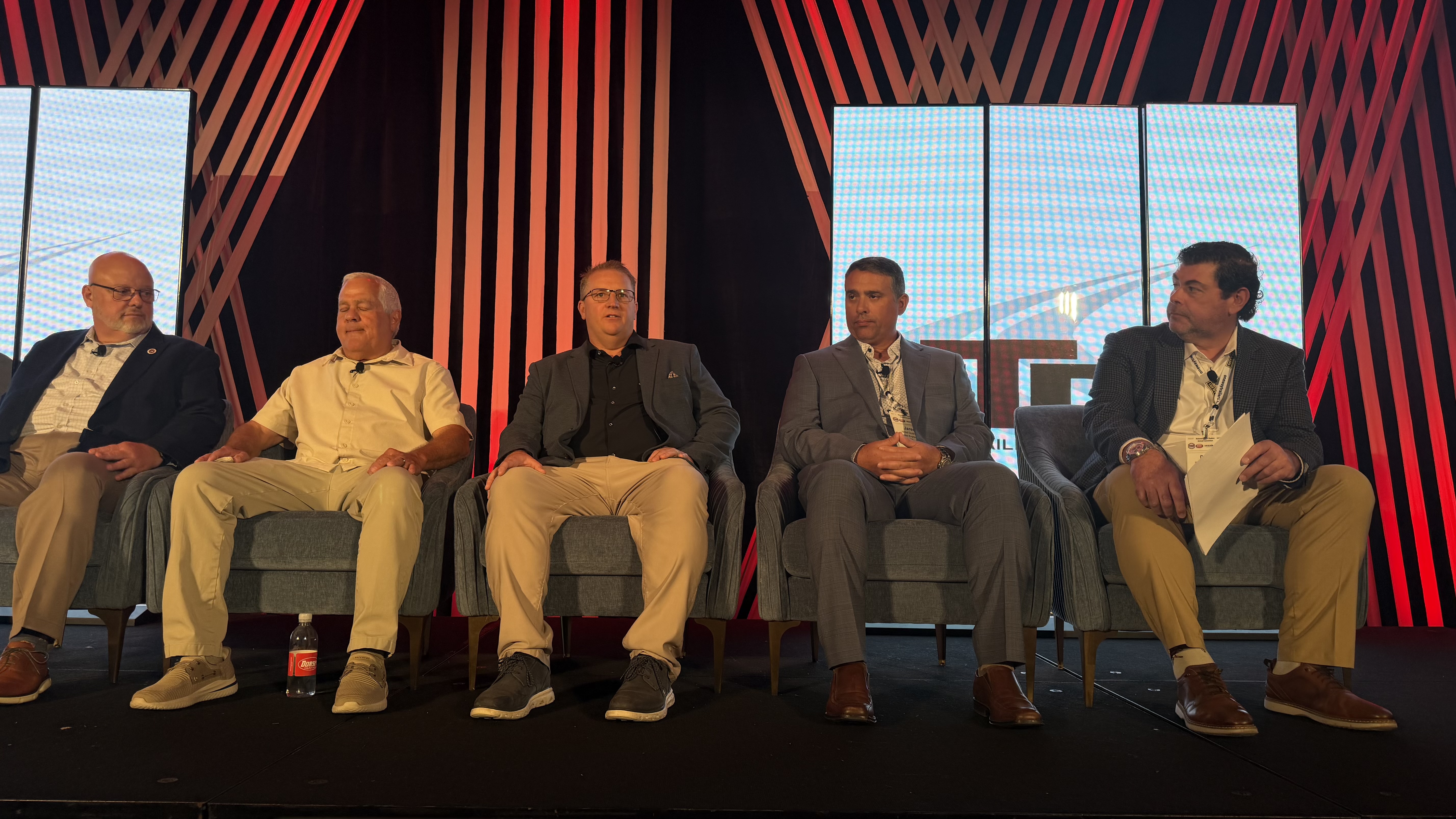 Five men seated on stage during a panel discussion with red striped backdrop and windows