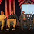 Five men seated on stage during a panel discussion with red striped backdrop and windows