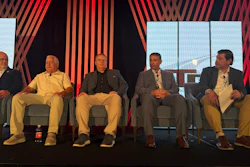 Five men seated on stage during a panel discussion with red striped backdrop and windows