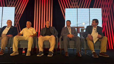 Five men seated on stage during a panel discussion with red striped backdrop and windows