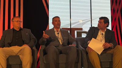 Three men in business attire participate in a panel discussion on stage with red striped backdrop and large windows.