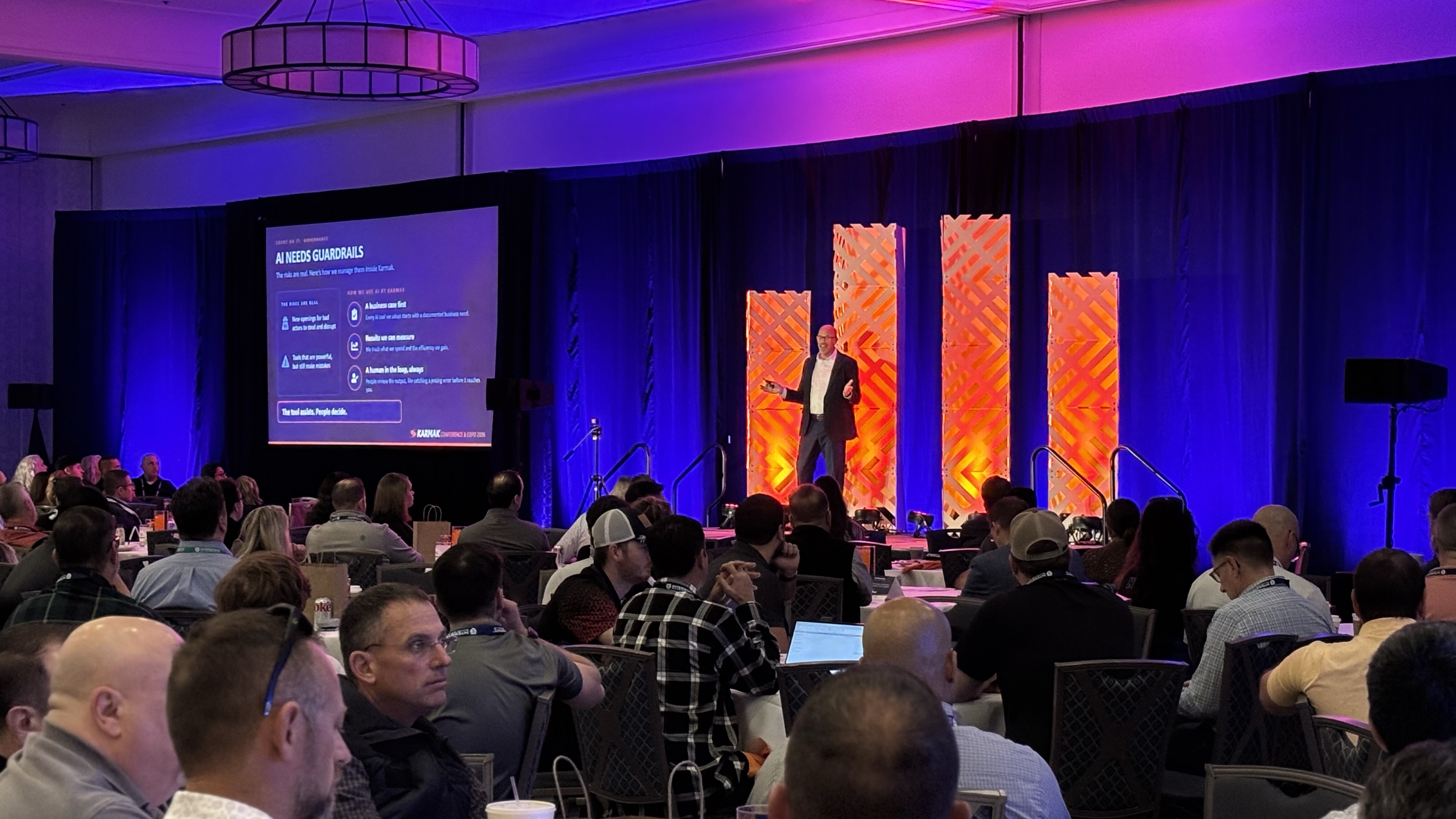 Conference audience seated at tables watching presenter on stage with purple and orange lighting and large display screens
