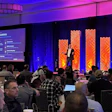 Conference audience seated at tables watching presenter on stage with purple and orange lighting and large display screens