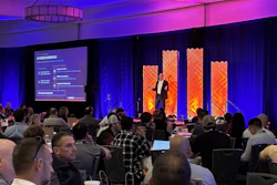 Conference audience seated at tables watching presenter on stage with purple and orange lighting and large display screens