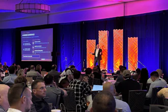 Conference audience seated at tables watching presenter on stage with purple and orange lighting and large display screens
