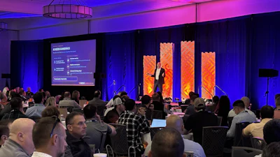 Conference audience seated at tables watching presenter on stage with purple and orange lighting and large display screens