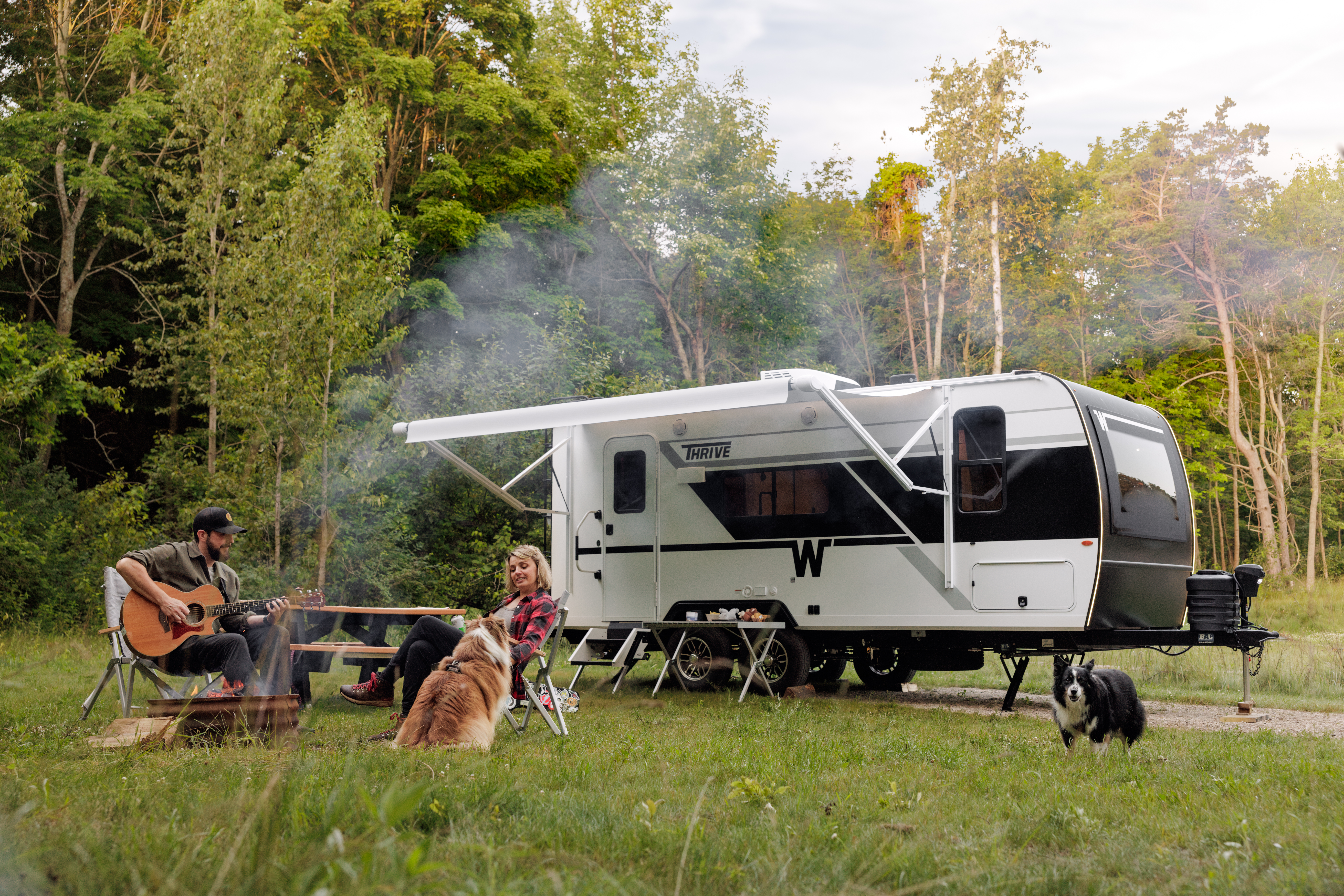 Family camping with travel trailer in forest clearing, man playing guitar while woman and child relax outdoors