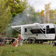 Family camping with travel trailer in forest clearing, man playing guitar while woman and child relax outdoors