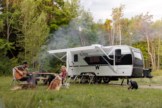 Family camping with travel trailer in forest clearing, man playing guitar while woman and child relax outdoors