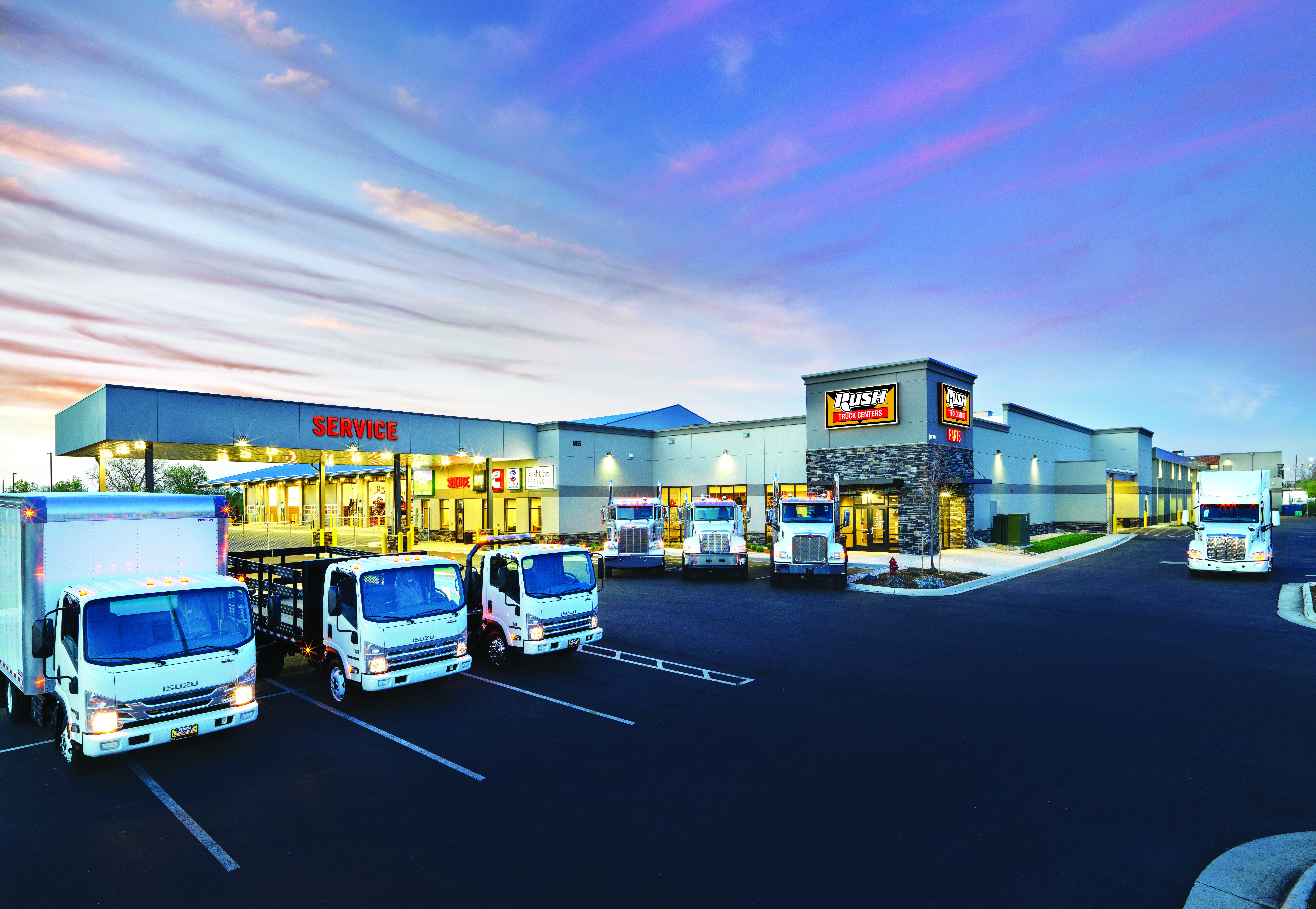 Isuzu commercial vehicle dealership facility at dusk with three white delivery trucks parked in front of modern building