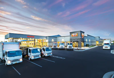 Isuzu commercial vehicle dealership facility at dusk with three white delivery trucks parked in front of modern building