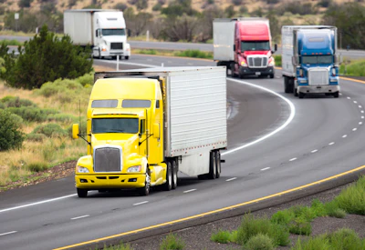 Yellow semi-truck leading convoy of commercial trucks on multi-lane highway through desert landscape