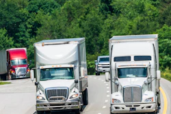 White commercial trucks driving on a multi-lane highway with dense green forest in the background