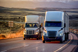 Two commercial semi-trucks driving on a highway through rolling hills and grassland during golden hour.