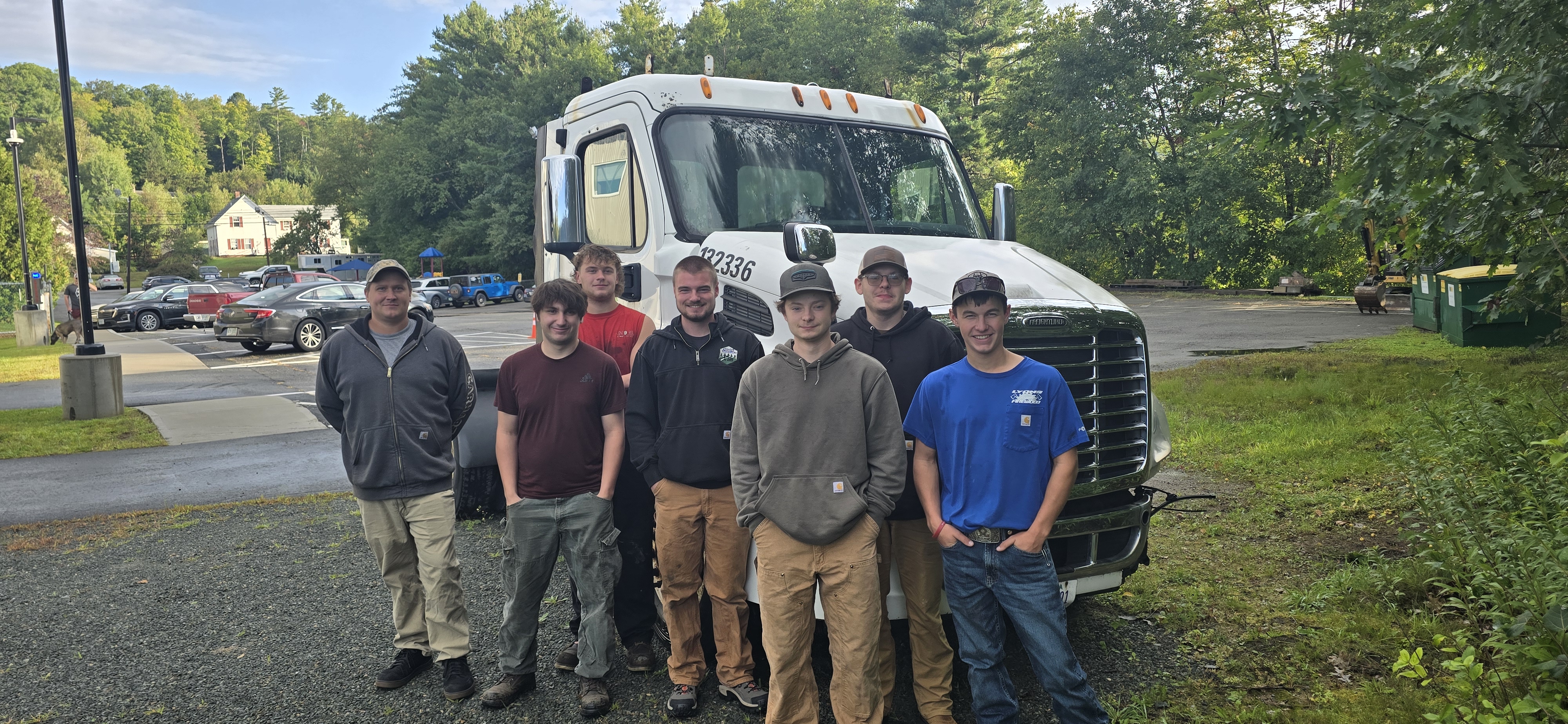 Seven workers pose together in front of a white commercial truck at a job site with trees and parked vehicles in background.