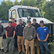 Seven workers pose together in front of a white commercial truck at a job site with trees and parked vehicles in background.