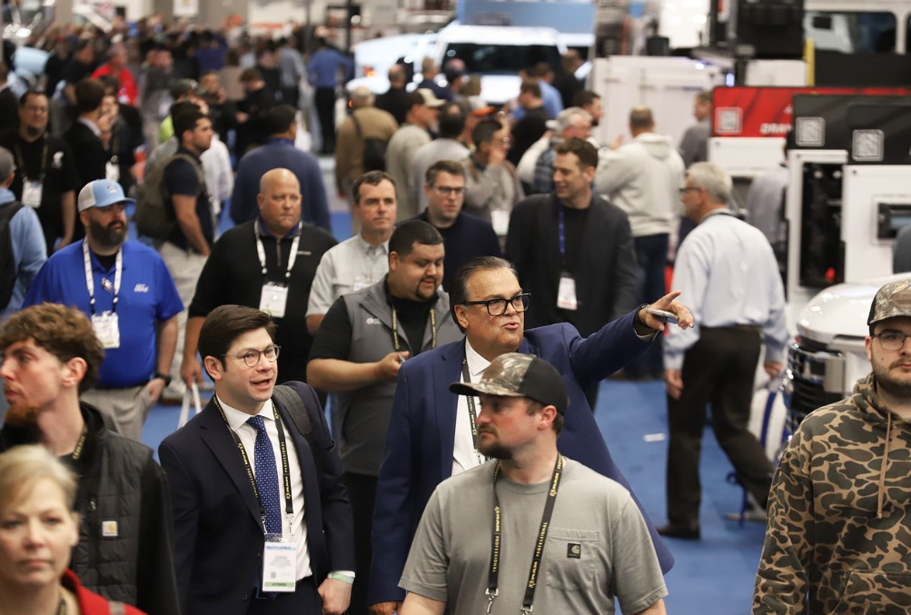 Crowded trade show floor with attendees wearing lanyards and badges networking among industrial equipment displays.