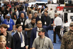 Crowded trade show floor with attendees wearing lanyards and badges networking among industrial equipment displays.