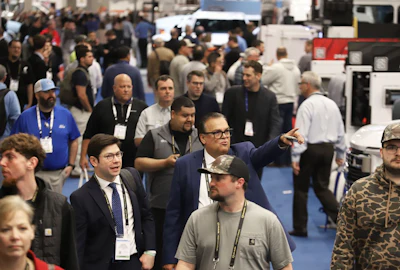 Crowded trade show floor with attendees wearing lanyards and badges networking among industrial equipment displays.